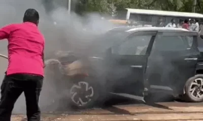 कार से उठती आग की लपटें और पाइप से आग बुझाने का प्रयास करते व्यक्ति।